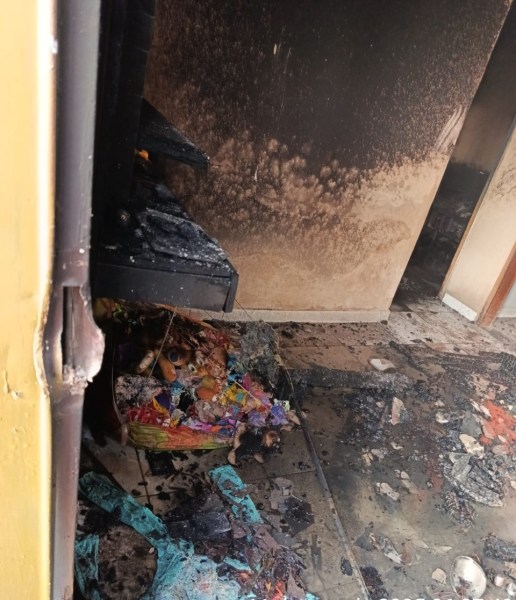 Interior de uma residência danificada por incêndio, com paredes carbonizadas e objetos queimados espalhados pelo chão.