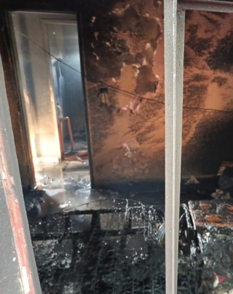 Visão do interior de uma residência que foi parcialmente destruída por um incêndio, mostrando paredes queimadas e móveis danificados.