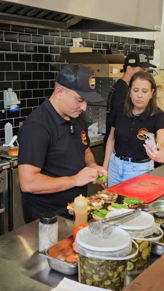 Um cozinheiro da Hamburgueria Novo Sabor preparando ingredientes com uma auxiliar em uma cozinha profissional.