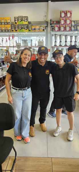 Três pessoas sorridentes vestindo camisetas e bonés da hamburgueria Novo Sabor, posando em um ambiente comercial com prateleiras ao fundo.