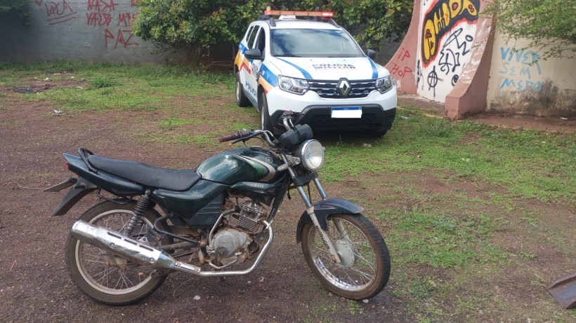 Motocicleta Yamaha YBR 125, cor verde, abandonada em terreno, com viatura da Polícia Militar ao fundo.