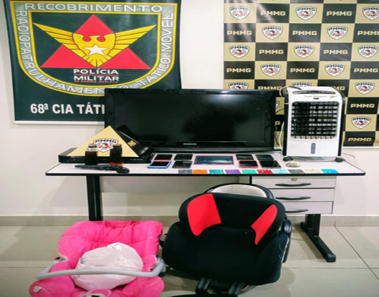 Mesa com diversos objetos apreendidos pela Polícia Militar, incluindo uma televisão, um ventilador, vários smartphones e um carrinho de bebê, com um fundo decorado com a bandeira e logotipos da Polícia Militar.