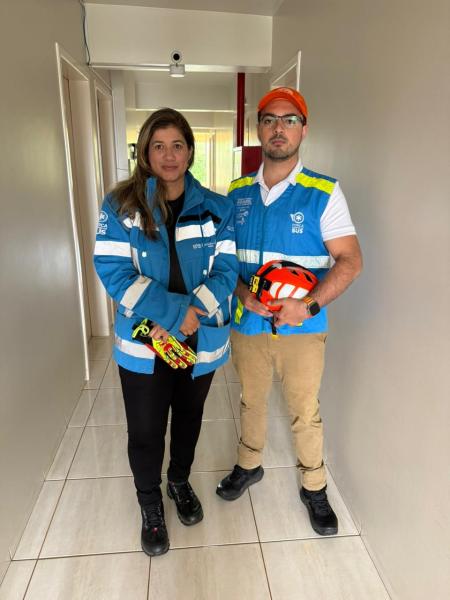 Dois profissionais de saúde, uma mulher e um homem, posando em um corredor. A mulher veste um casaco azul e o homem usa um colete azul e um boné laranja, ambos segurando equipamentos de emergência.