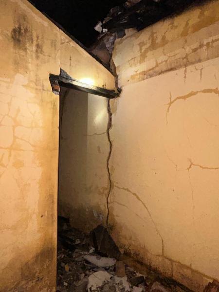 Interior de um imóvel abandonado com paredes danificadas e marcas de incêndio, mostrando uma rachadura significativa e detritos no chão.
