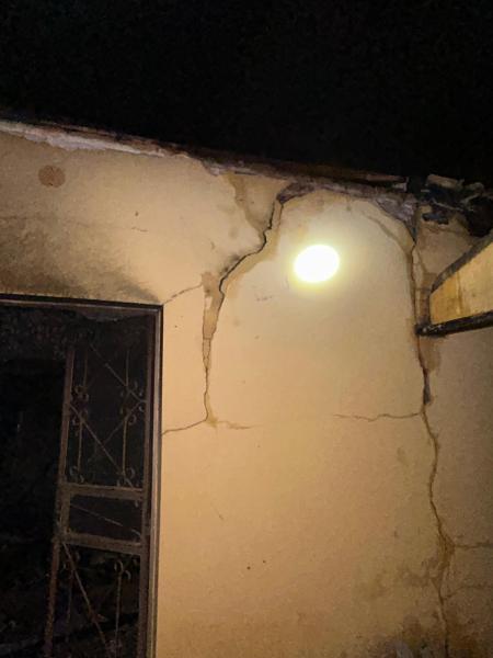 Imagem de uma parede de imóvel abandonado com rachaduras visíveis, um ponto de luz iluminando a área. A estrutura parece apresentar danos significativos.