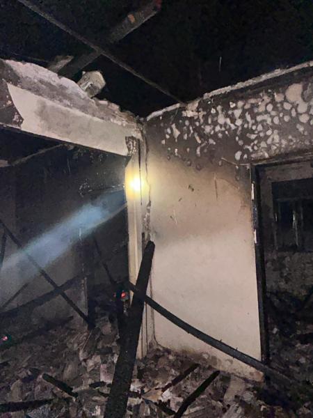 Interior de um imóvel abandonado com paredes danificadas pelo fogo, colunas pretas e destroços espalhados pelo chão, iluminado por um feixe de luz.