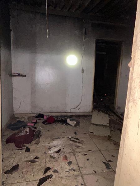 Interior de um imóvel abandonado com paredes sujas e piso coberto de detritos. Um ponto de luz pode ser visto na parede, indicando uma fonte de iluminação. O ambiente aparenta estar em estado de deterioração.