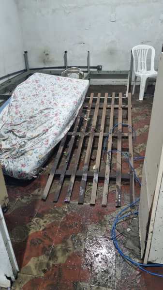 Interior de uma residência com piso molhado e madeiramento danificado, mostrando um colchão e um estrado de cama, além de uma cadeira plástica.