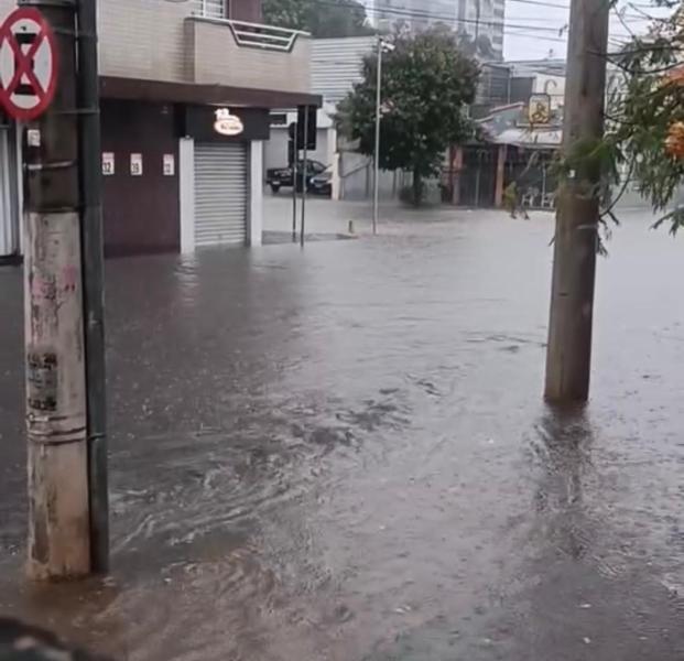 Cenário de alagamento em uma rua com água cobrindo a calçada e parte dos edifícios ao fundo, com um poste e placas de trânsito visíveis.