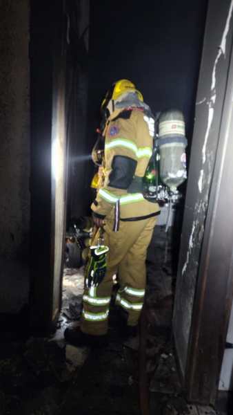 Um bombeiro em ação em uma residência danificada por incêndio, usando equipamento de proteção e respirador, enquanto avalia a situação interna.