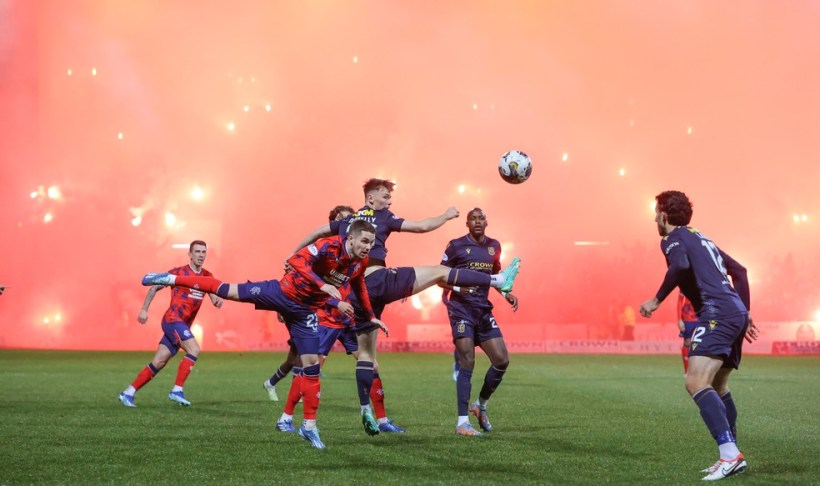 Jogadores de futebol em ação durante um jogo, com um jogador fazendo um chute acrobático em meio a uma intensa fumaça e fogos de artifício ao fundo.
