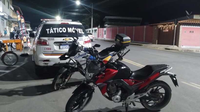 Motocicleta preta estacionada ao lado de uma viatura da Polícia Militar em via pública à noite, com letreiro 'TÁTICO MÓVEL' visível.