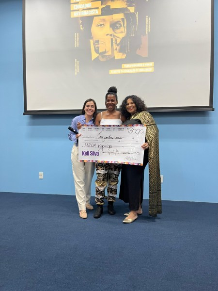 Cerimônia do Prêmio Educador Antirracista em Divinópolis com três mulheres segurando um cheque simbólico de R$ 300,00, reconhecendo o projeto H2OR Hip Hop. Ao fundo, um banner com informações sobre o prêmio.