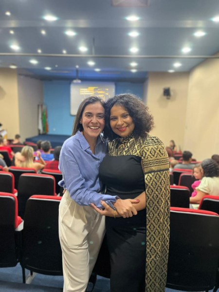 Duas mulheres sorrindo e se abraçando em um auditório, com cadeiras vermelhas ao fundo e uma apresentação visível na tela.