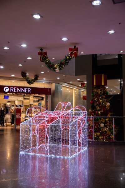 Decoração natalina com um grande presente iluminado em um shopping, adornado com laços vermelhos e luzes, rodeado por outras decorações festivas.