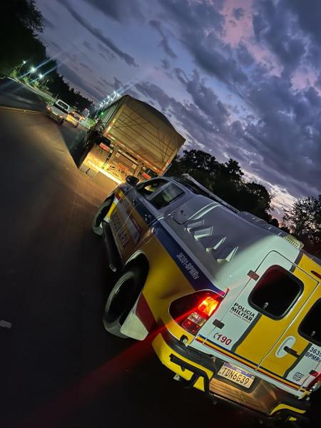 Viatura da Polícia Militar Rodoviária parada em uma rodovia à noite, com caminhão carregado visível à frente e céu com nuvens ao fundo.