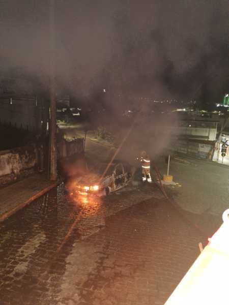 Um carro em chamas sendo combatido por bombeiros em uma rua à noite, com fumaça saindo do veículo danificado.
