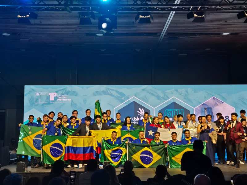 Participantes da 6ª edição da WorldSkills Américas posam em grupo no evento, segurando bandeiras do Brasil e de outros países, em Santiago, Chile.