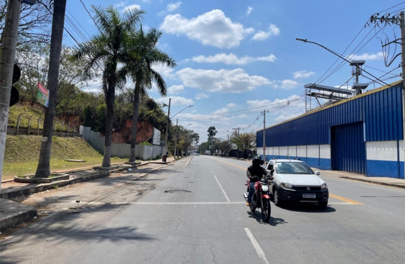 Vista da Avenida Autorama em Divinópolis, mostrando uma rua com um motociclista e um carro, cercada por árvores e postes, com um céu parcialmente nublado.