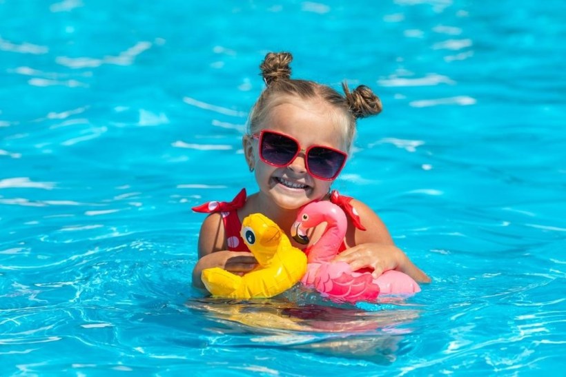 Criança sorridente em uma piscina, segurando boias de pato e flamingo, vestindo óculos de sol.