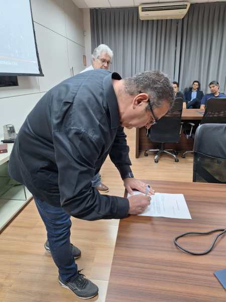 Homem assinando um documento em uma mesa, com outras pessoas ao fundo em um ambiente de escritório.