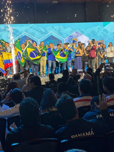 Grupo de competidores brasileiros celebrando com bandeiras do Brasil em um evento, cercados por fogos de artifício e uma plateia animada.