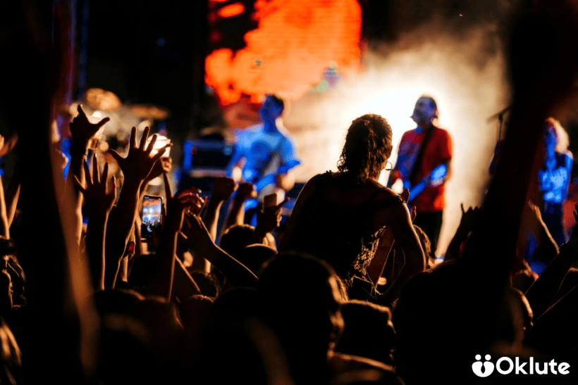 Pessoas em um show ao vivo, com muitas mãos levantadas, enquanto uma banda toca ao fundo, iluminada por luzes coloridas.