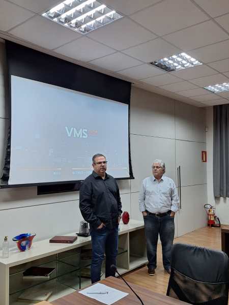 Homens em pé durante uma apresentação, com um projetor exibindo uma apresentação ao fundo. Um deles fala enquanto o outro observa.