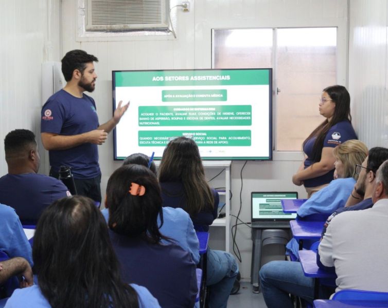 Um instrutor apresenta um slide sobre assistência e serviços em uma sala de treinamento, enquanto os participantes assistem atentamente.