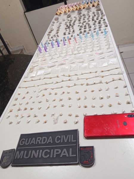 Mesa com drogas apreendidas, incluindo porções de maconha, cocaína e crack, além de um celular e distintivos da Guarda Civil Municipal.