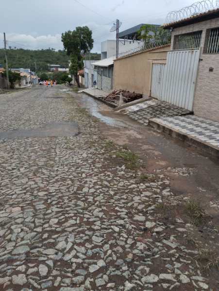 Rua com pavimentação irregular e água acumulada, com alguns restos de materiais de construção visíveis, cercada por propriedades em Divinópolis.