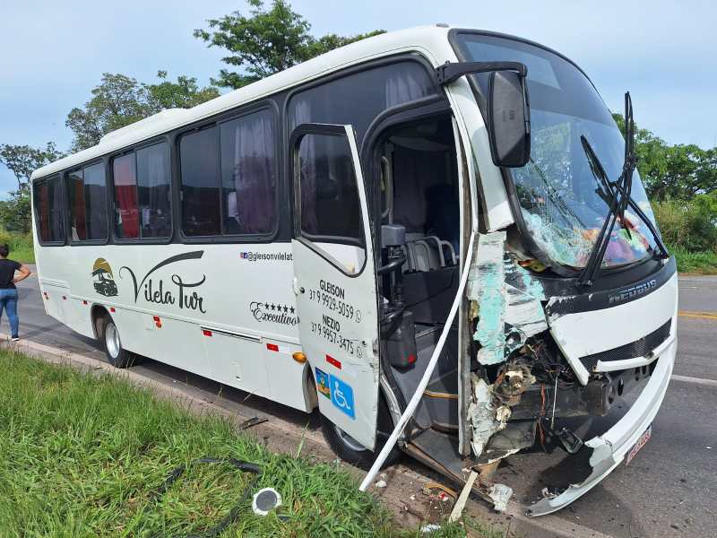 Um ônibus de turismo branco com o nome 'Viela Tur' está danificado, com a parte frontal colidida. A cena mostra a estrada e vegetação ao fundo, com uma pessoa ao lado do ônibus.