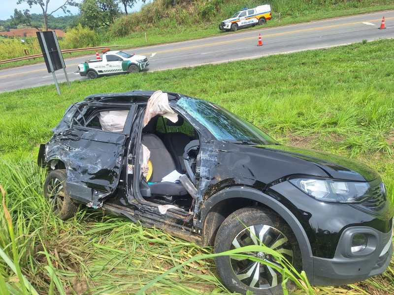 Um carro preto danificado, com uma porta aberta, está parado em uma área gramada próximo a uma estrada. Um veículo da polícia é visível ao fundo.