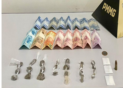 Vários pacotes de substâncias aparentando ser drogas, acompanhados por notas de dinheiro e uma cápsula de munição, dispostos sobre uma mesa com um pequeno marcador da Polícia Militar de Minas Gerais ao fundo.