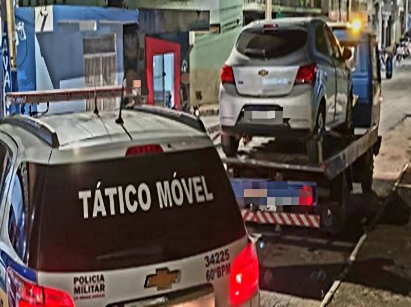 Veículo sendo rebocado por uma caminhonete da Polícia Militar em uma rua à noite, com outros veículos e construções ao fundo.