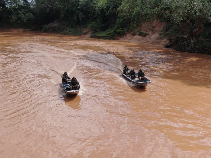 Dois barcos da Polícia Militar navegando em um rio de águas marrons, com membros da equipe a bordo, cercados por vegetação ao longo das margens.