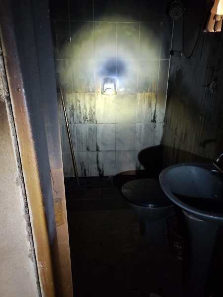 Interior de banheiro com marcas de fumaça nas paredes e um banheiro danificado após incêndio, iluminação fraca com luz do lado de fora.