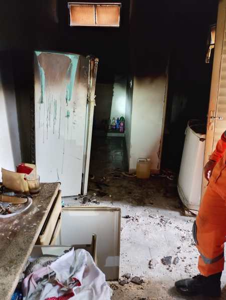 Imagem da cozinha de uma residência afetada por um incêndio, com paredes e móveis danificados por fumaça e fogo. Um frigorífico sujo é visível, e o chão está coberto de detritos e roupas. Um bombeiro, vestido com uniforme de proteção, está presente no local.