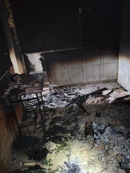 Interior de um quarto danificado por um incêndio, com paredes pretas e chão coberto de cinzas e destroços.