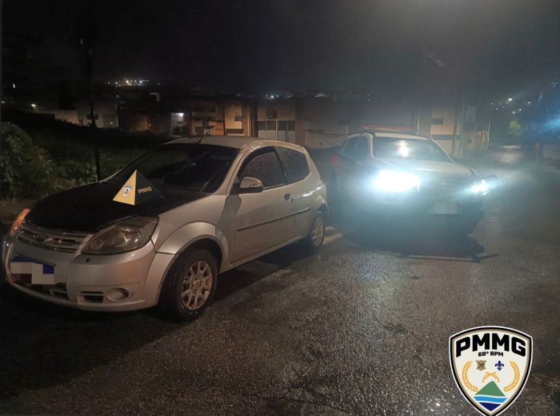 Imagem de um carro branco estacionado à noite, próximo a um veículo da polícia com faróis acesos, em uma área urbana de Nova Serrana.