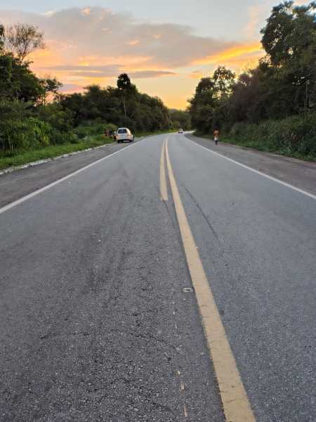A view of a deserted road bordered by trees and greenery, with gentle hills and a sunset in the background. A car is parked on the side of the road, and a person is walking in the distance.