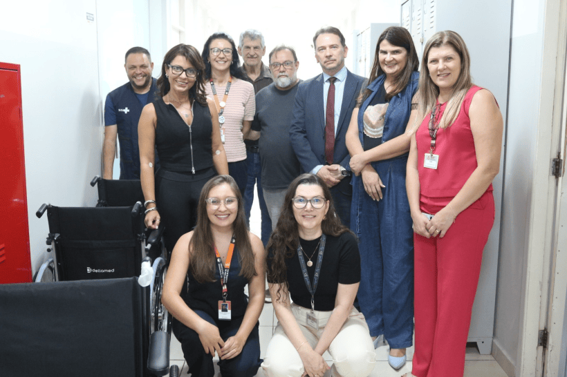 Grupo de pessoas posando em uma unidade de saúde, com cadeiras de rodas ao fundo, durante a entrega de doações.