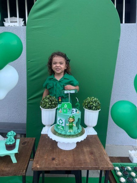 Uma garota sorridente vestindo uma camisa verde ao lado de um bolo de aniversário decorado com personagens e elementos em verde, com balões verdes e brancos ao fundo.