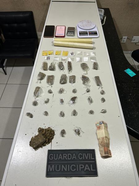 Mesa com substâncias ilícitas apreendidas, incluindo maconha, cocaína e ecstasy, além de dinheiro, balanças de precisão e materiais de acondicionamento. Placa da Guarda Civil Municipal em destaque.