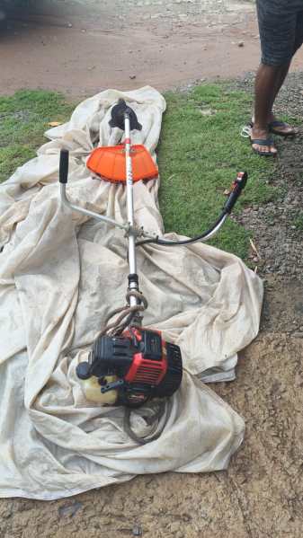 A gas-powered string trimmer lying on a tarp on the ground, with an orange guard and black engine, with a person partially visible in the background.