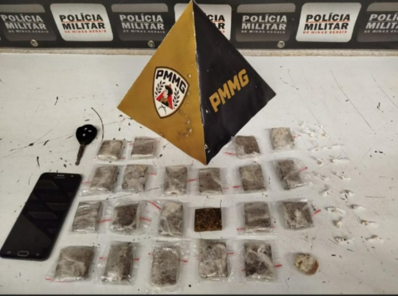 Imagem de uma mesa com drogas apreendidas, incluindo pacotes de maconha e pedras de crack, junto a um telefone celular e um objeto em forma de pirâmide com o logotipo da PMMG ao fundo.