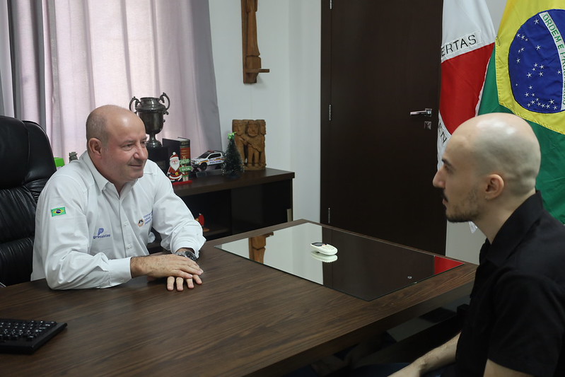 Reunião entre o Presidente da Câmara Municipal de Divinópolis e um novo servidor em um escritório, com bandeira do Brasil ao fundo.