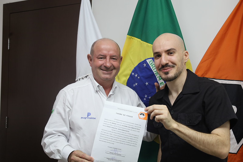 O Presidente da Câmara Municipal de Divinópolis, Israel da Farmácia, e o novo servidor Guilherme Klein Camara de Paula, segurando o termo de posse, em frente a bandeiras do Brasil e de Divinópolis.