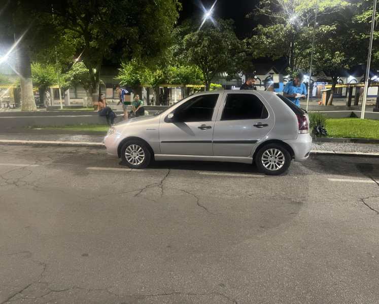 Carro prata estacionado em via pública, com agentes da Guarda Civil Municipal fazendo abordagens ao fundo, em um ambiente urbano noturno.