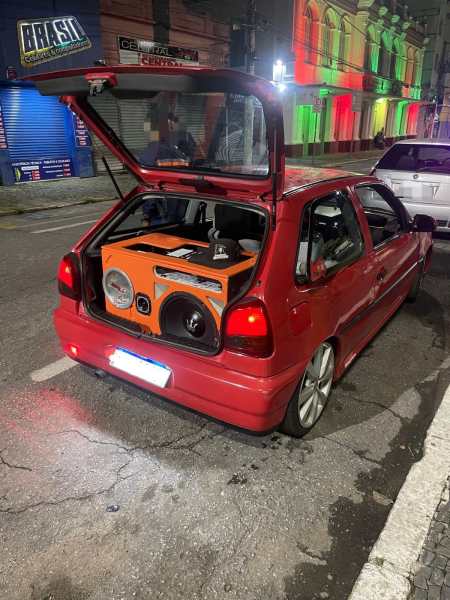 Carro vermelho com porta-malas aberto, exibindo um sistema de som potente, em uma rua iluminada à noite.